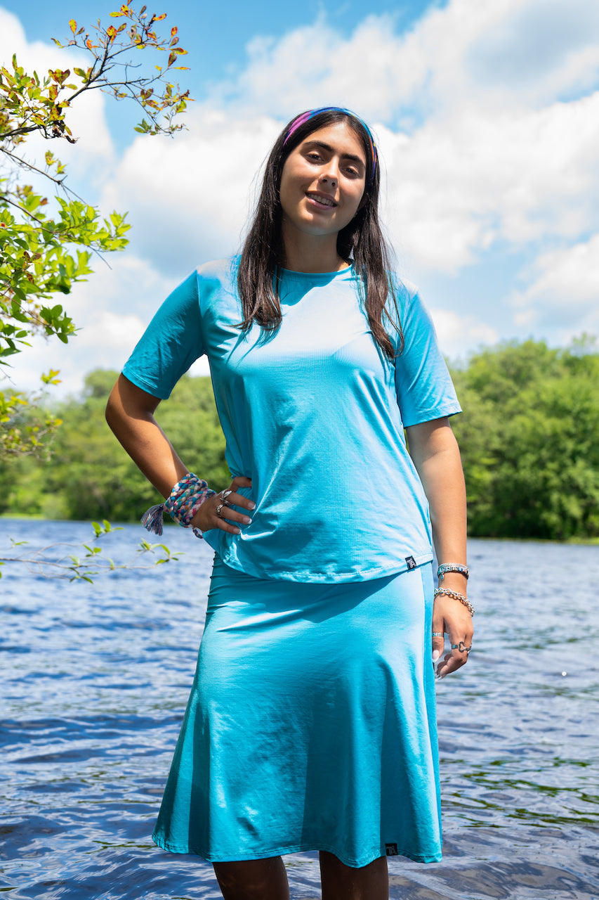 Woman in a blue outfit standing by a body of water with trees and clouds in the background