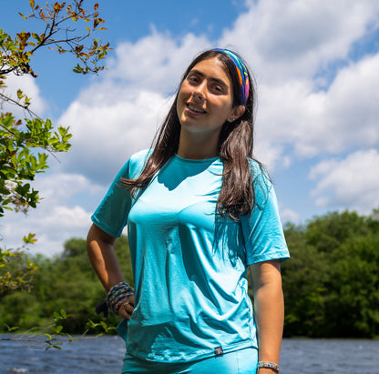 Woman in a turquoise shirt standing outdoors with trees and water in the background