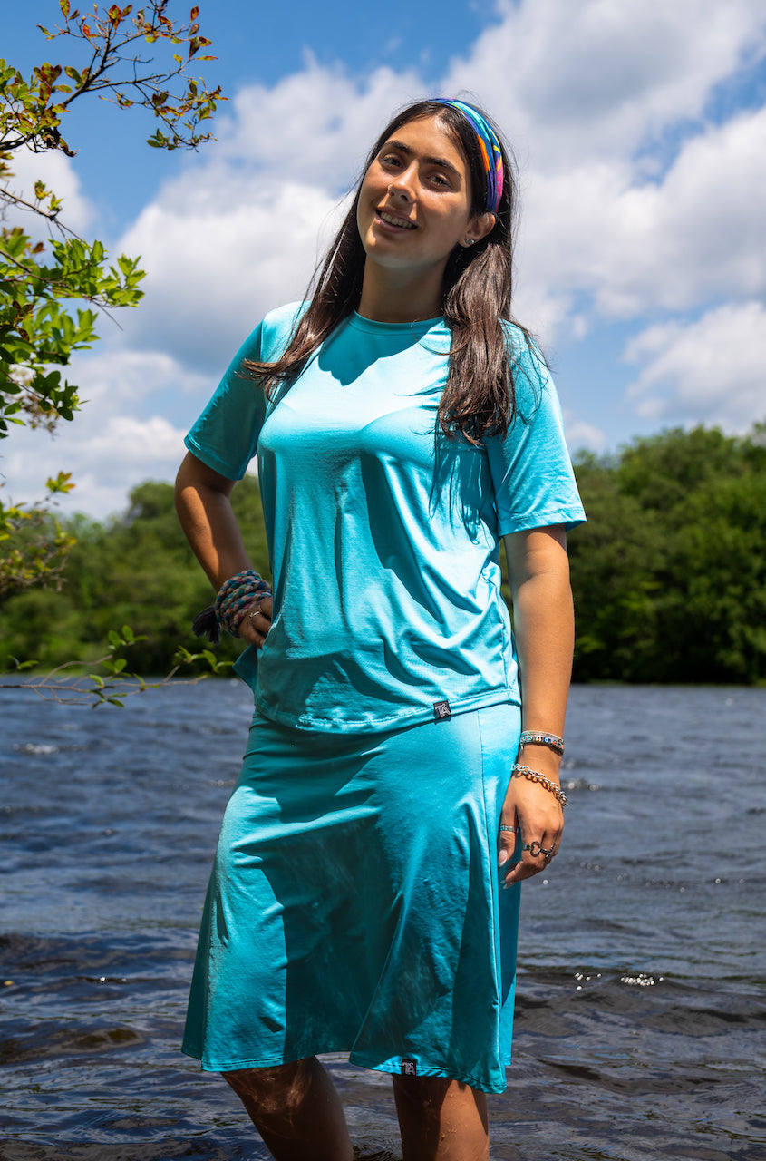 Woman in a turquoise dress standing by a body of water with trees and blue sky in the background