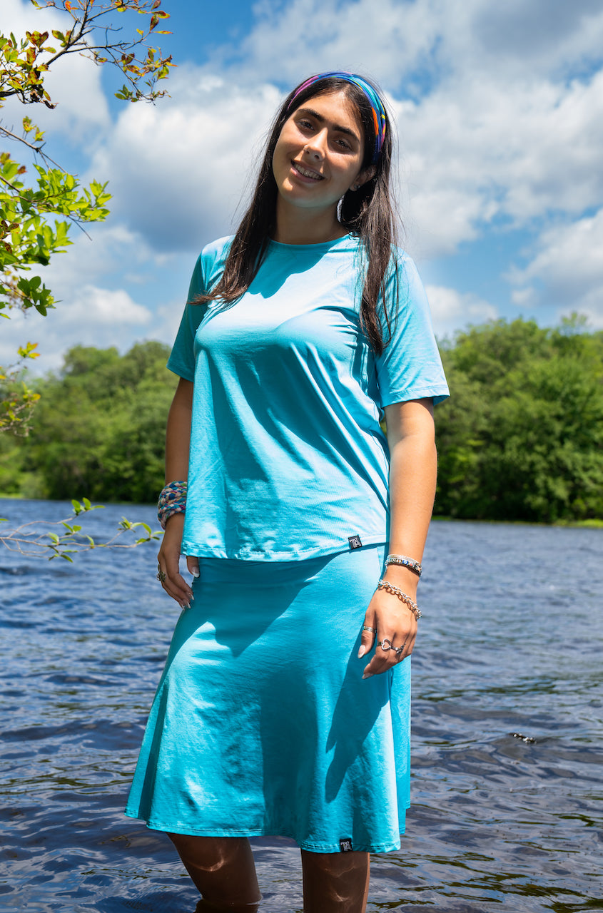 Woman wearing a blue dress standing by a body of water with trees and clouds in the background